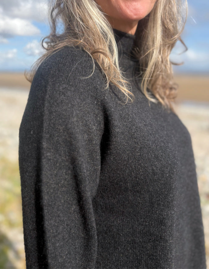 Person wearing a black yak wool sweater and blue jeans standing on a beach with a scenic background.