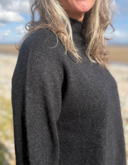 Person wearing a black yak wool sweater and blue jeans standing on a beach with a scenic background.