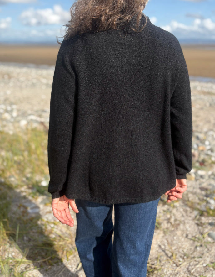 Person wearing a black yak wool sweater and blue jeans standing on a beach with a scenic background.