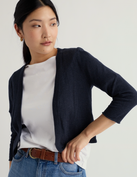 Woman wearing a navy cardigan over a white shirt with blue jeans on a plain background