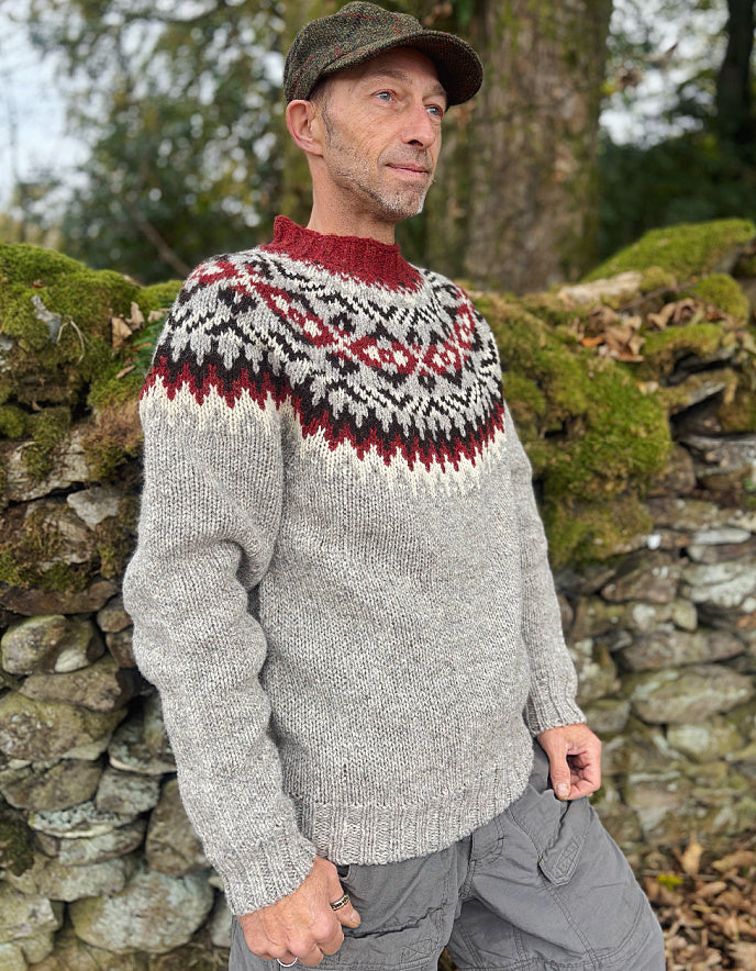 Man wearing a patterned sweater standing against a stone wall with greenery.