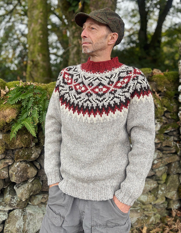 Man wearing a patterned sweater and cap standing against a stone wall with greenery.