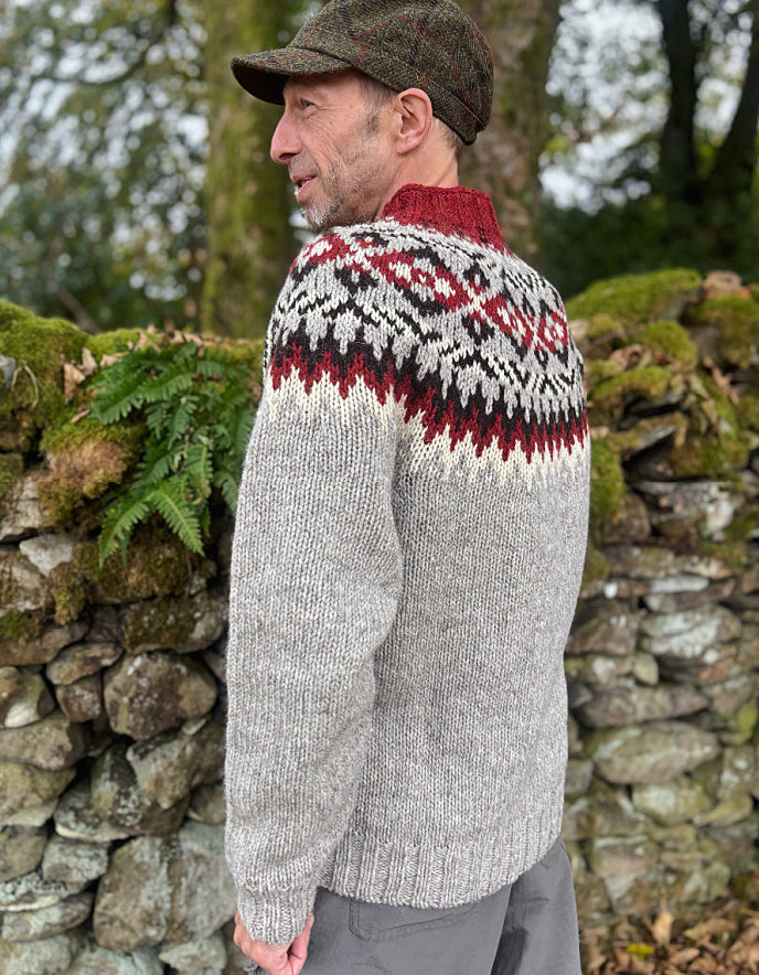 Man wearing a patterned sweater and cap standing against a stone wall with greenery.