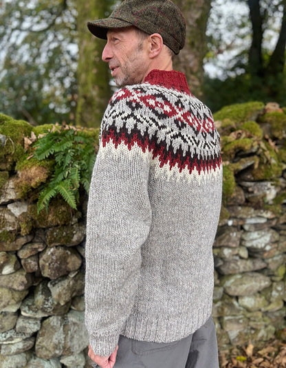 Man wearing a patterned sweater and cap standing against a stone wall with greenery.