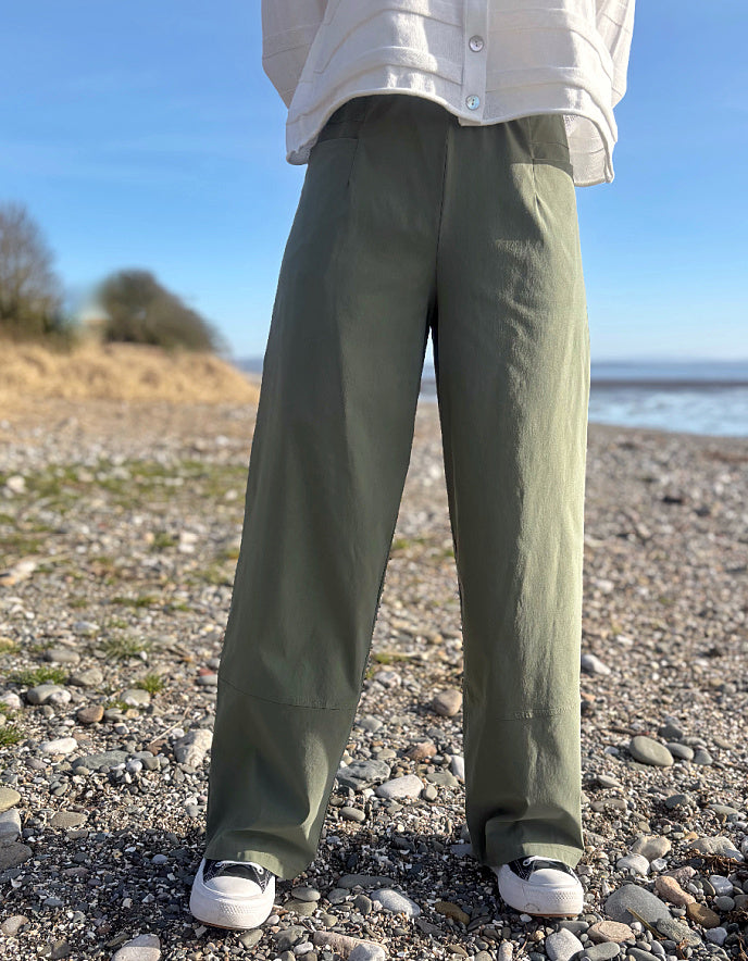 Person wearing green pants and a white shirt on a pebbly beach with ocean view