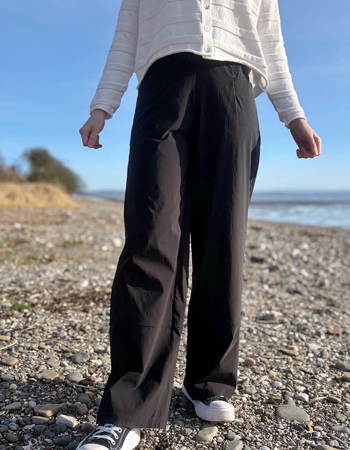 Person wearing black pants and a white shirt standing on a pebbly beach with a clear blue sky.