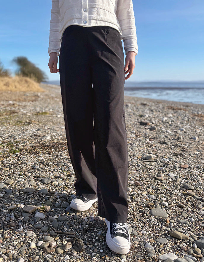 Person wearing black pants and white sneakers on a pebbly beach with a blue sky.