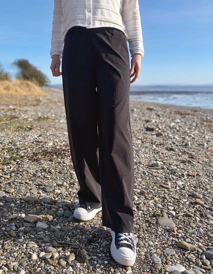 Person wearing black pants and white sneakers on a pebbly beach with a blue sky.