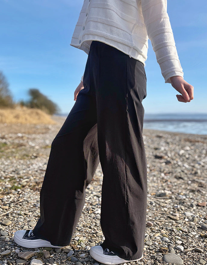 Person wearing black pants and a white jacket standing on a pebbly beach with a clear sky.