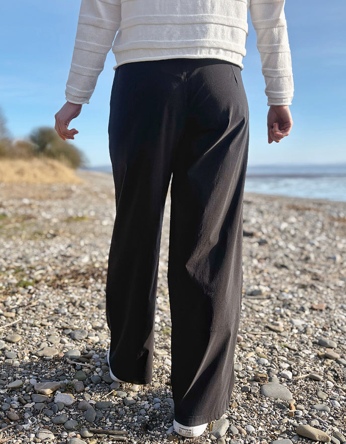 Person wearing black pants and a white jacket standing on a pebbly beach with ocean and sky in the background.