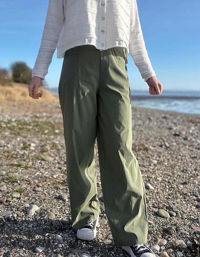 Person wearing green pants and a white jacket standing on a pebbly beach with a clear sky.
