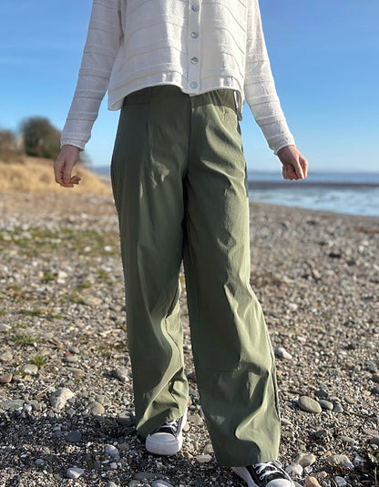 Person wearing green pants and a white jacket standing on a pebbly beach with a clear sky.