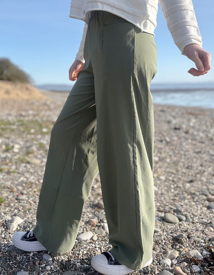 Person wearing green pants and white sneakers standing on a pebbly beach with a clear sky.