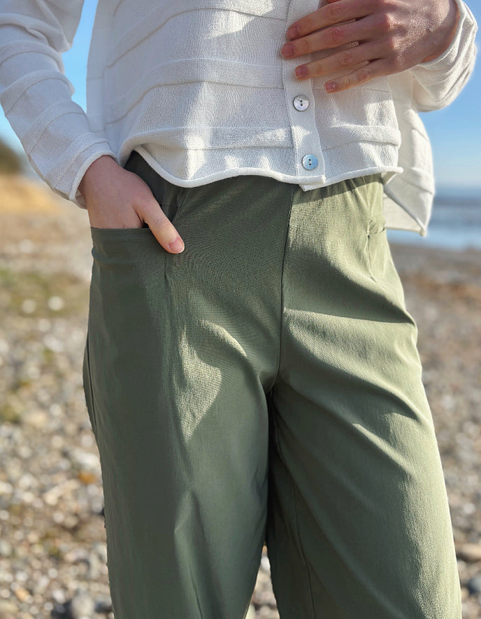 Person wearing a white shirt and green pants standing on a pebbly beach.