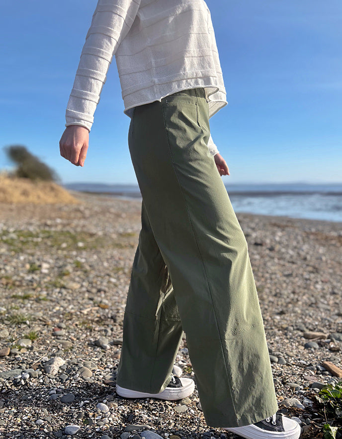 Person wearing green pants and a white shirt on a pebbly beach with ocean view