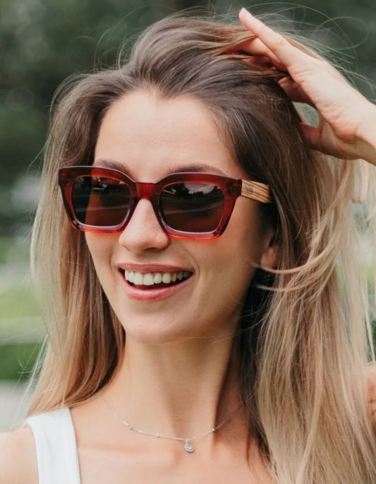 A woman with blonde hair smiling outdoors while wearing chunky square sunglasses with a translucent reddish-brown frame and light wood temple tips.