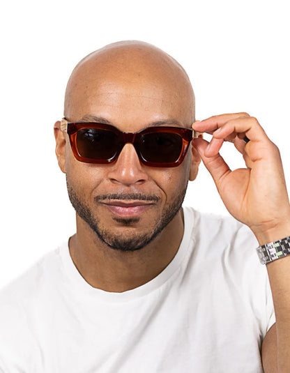 A man wearing dark brown acetate square sunglasses with dark lenses, holding one side of the frame with his hand against a plain white background.
