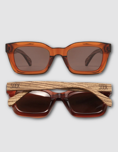 Two pairs of sunglasses shown on a light gray background: one with translucent amber-orange frames and brown lenses, and one with wood-grain arms and dark brown lenses. The wood arms are branded with 'SOCK'.