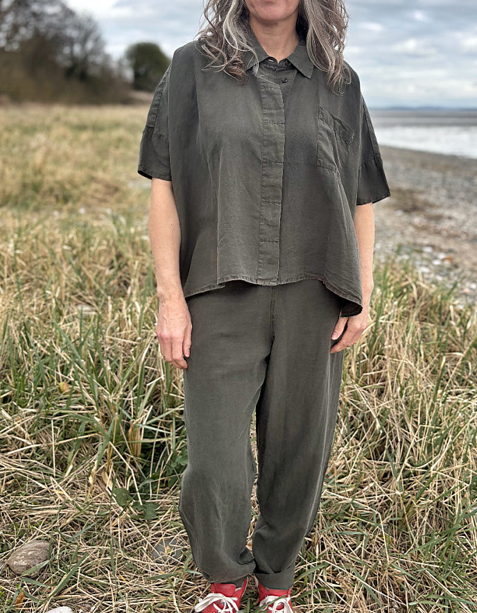 Person standing in a field wearing a green tencel oversized shirt, white pants, and red sneakers.