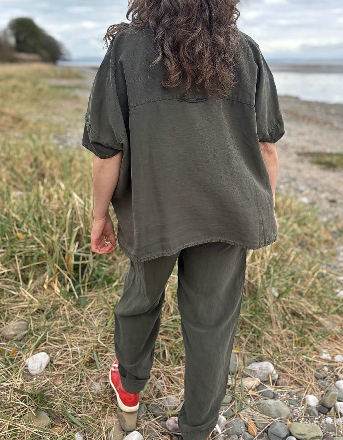 Person standing in a field wearing a green tencel oversized shirt, white pants, and red sneakers.