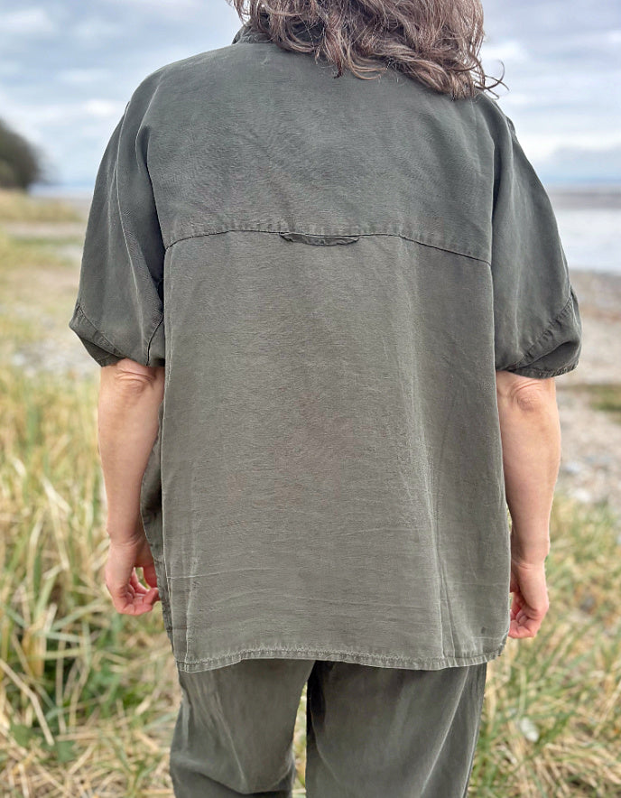 Person standing in a field wearing a green tencel oversized shirt, white pants, and red sneakers.