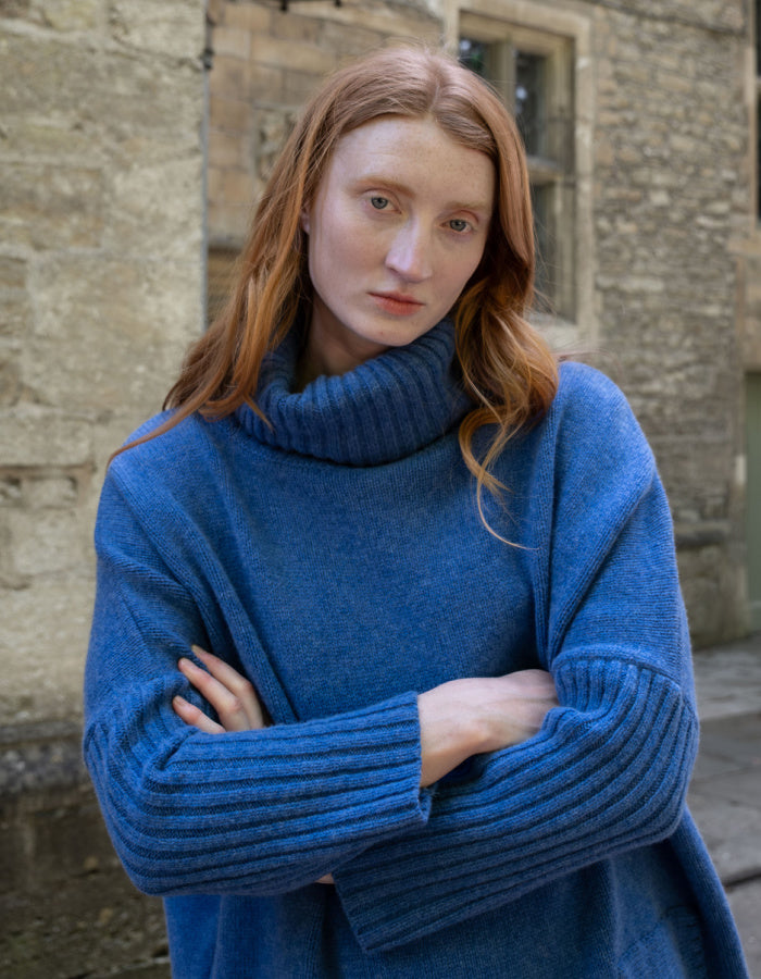 A woman with long red hair wearing a chunky blue ribbed turtleneck sweater, standing with arms crossed in front of a stone building.