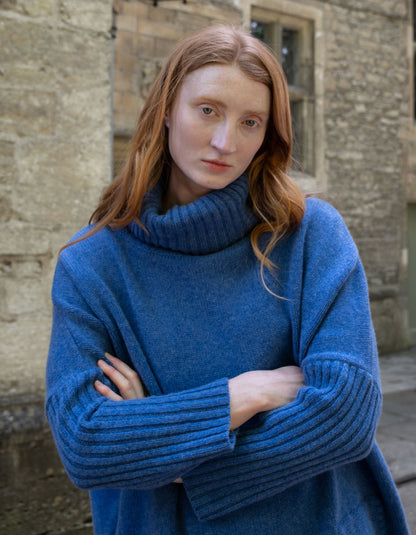 A woman with long red hair wearing a chunky blue ribbed turtleneck sweater, standing with arms crossed in front of a stone building.