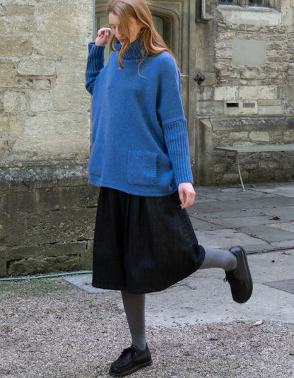 A woman wearing a blue turtleneck knit sweater with ribbed elbow patches, a black midi skirt, gray tights, and black shoes, standing outdoors in front of a stone building.