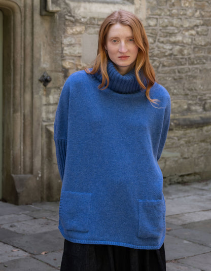 A woman with red hair wearing an oversized blue turtleneck sweater with front patch pockets, standing outdoors against a stone wall.