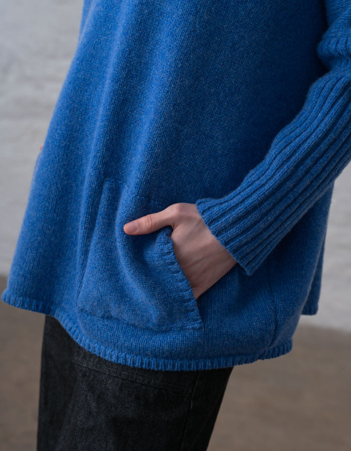A close-up of a person wearing a textured blue knit sweater with a side pocket, shown from the chest down with a hand tucked into the pocket.