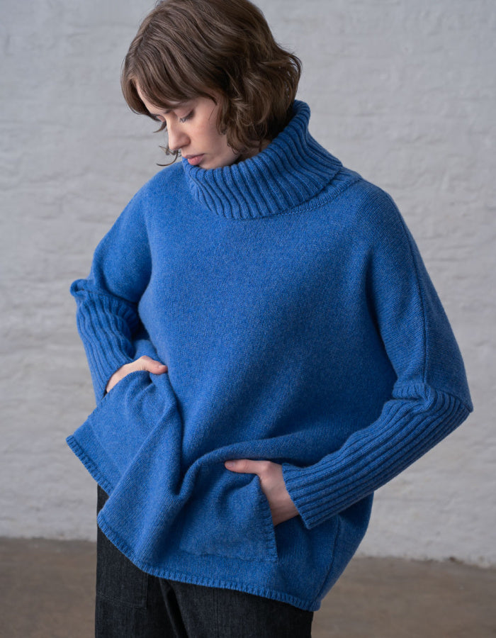 A woman wearing a vibrant blue turtleneck sweater with ribbed cuffs and hem, standing against a light gray textured wall.