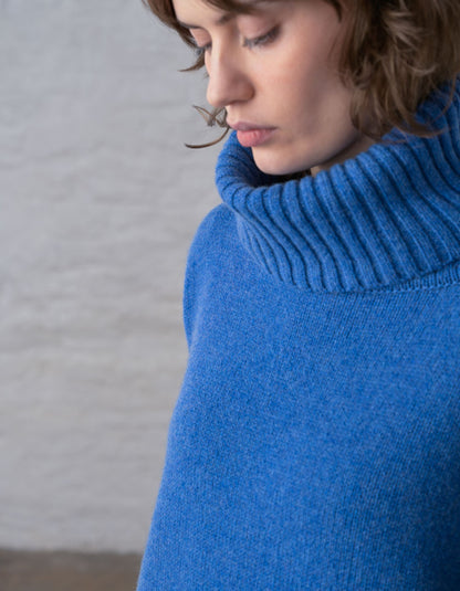 A close-up of a woman wearing a bright blue ribbed turtleneck sweater, with her face partially visible and looking downward against a soft light gray background.