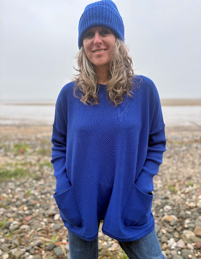 A woman wearing a vibrant royal blue knit sweater and matching blue beanie stands on a pebble beach with a cloudy sky in the background.