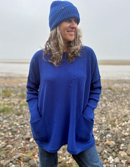 A woman wearing a vibrant royal blue knit sweater with a wavy textured pattern, a matching blue beanie, and blue jeans, standing on a pebble beach under an overcast sky.