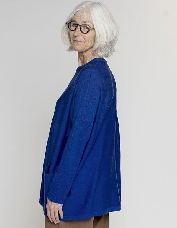 An older woman with short white hair and glasses, wearing a textured royal blue long-sleeve top, shown from a side-back profile against a light gray studio background.