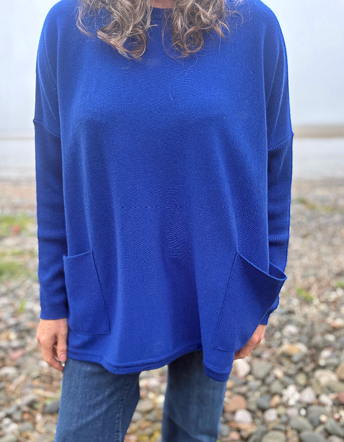 A person wearing a vibrant royal blue long-sleeve knit sweater with front patch pockets, paired with blue jeans, standing outdoors on a pebble beach.