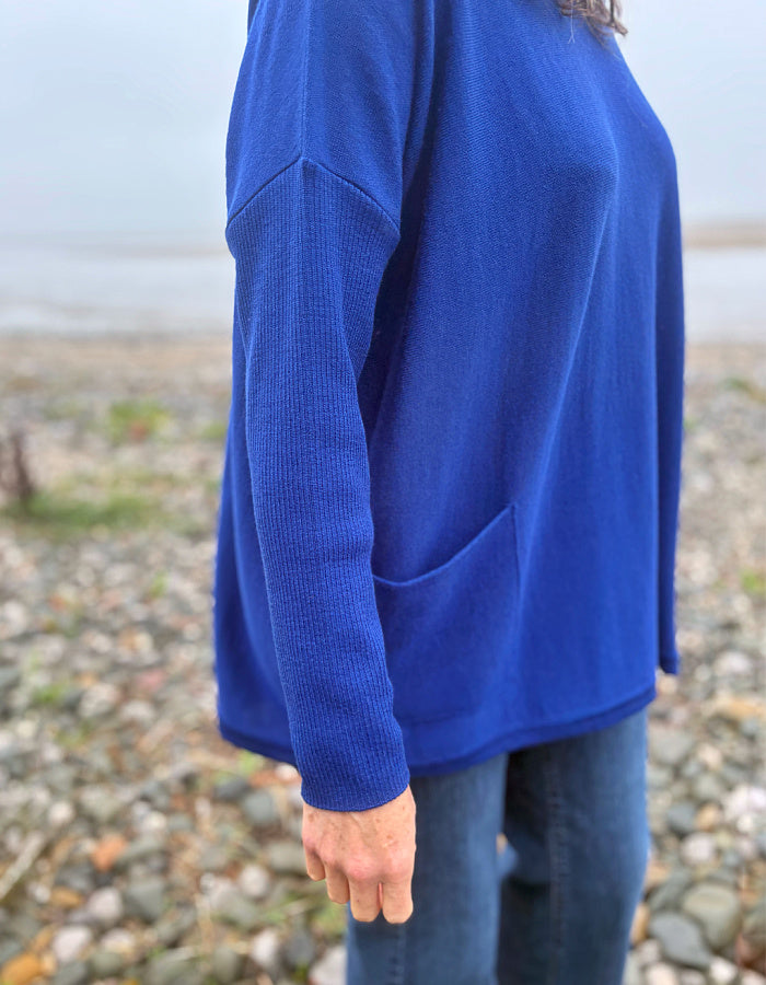 A person wearing a bright royal blue knit sweater with a front patch pocket, standing on a pebble beach.