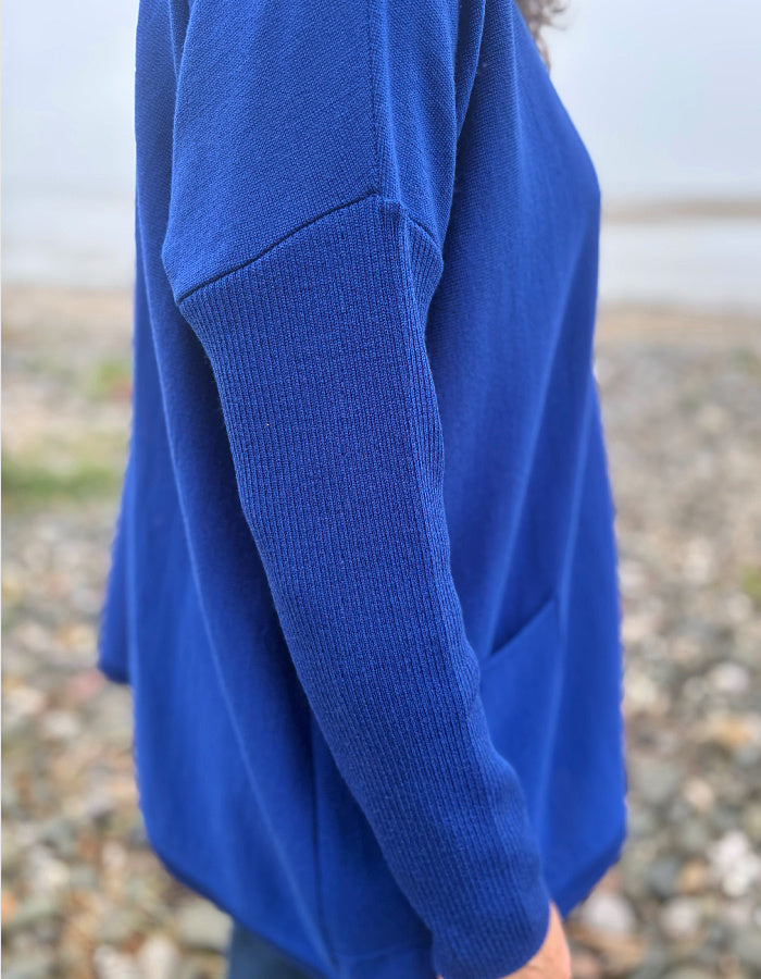 A close-up side view of a person wearing a vibrant royal blue knit sweater, standing on a pebble beach with a blurred background.