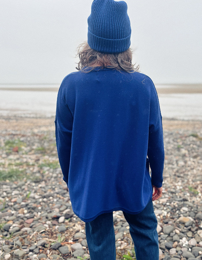 A person standing on a pebble beach, viewed from behind, wearing a dark blue knit beanie, a dark blue long-sleeve top, and dark blue pants.
