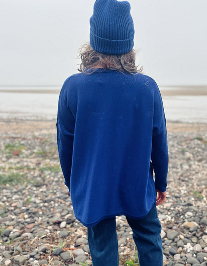 A person standing on a pebble beach, viewed from behind, wearing a dark blue knit beanie, a dark blue long-sleeve top, and dark blue pants.