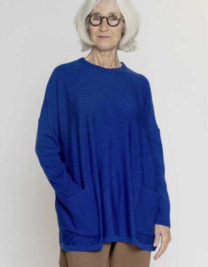 An older woman with short white hair and glasses wearing a vibrant royal blue knit sweater with side pockets, standing against a plain light gray background.