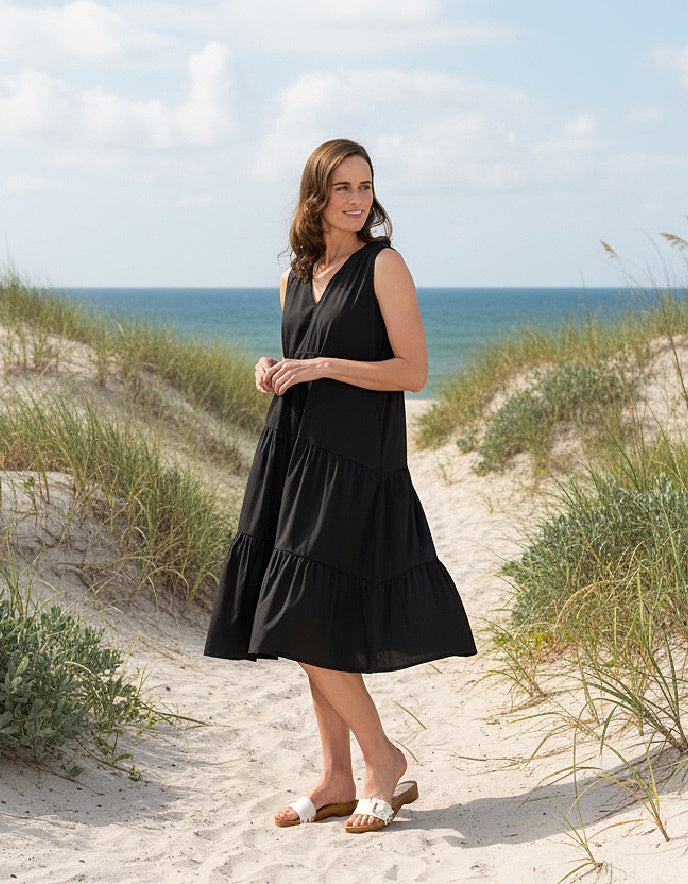 Woman in a black dress standing on a sandy beach with grass and ocean in the background