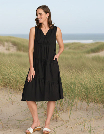 Woman in a black dress standing on a sandy beach with grasses.