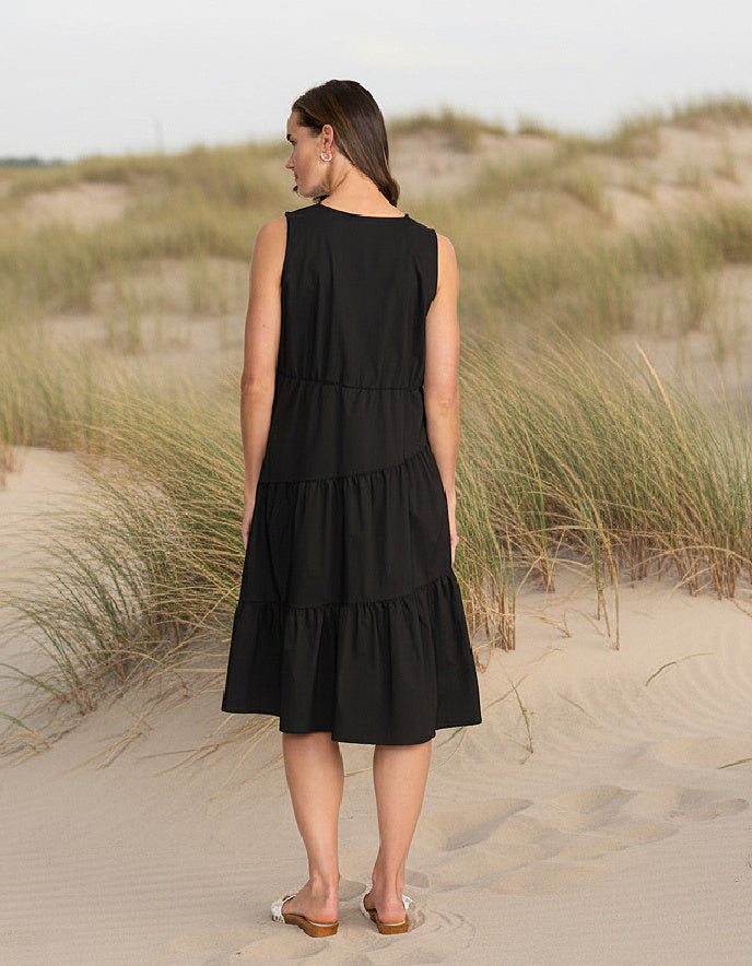 Woman in a black dress standing on a sandy dune with grasses.