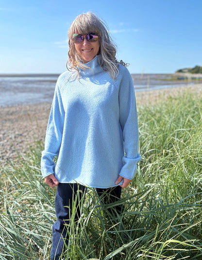 Person wearing a light blue sweater standing in tall grass by a beach.