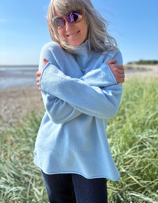 Person wearing a light blue sweater standing on a beach with grass and water in the background