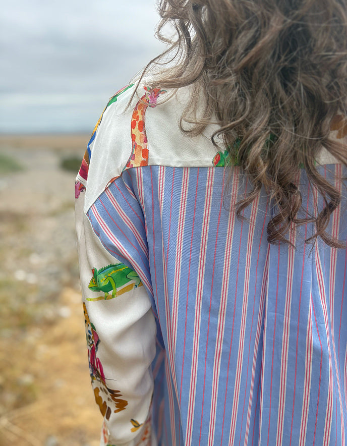 A close-up back view of a person wearing a long-sleeve shirt with a blue and red vertical stripe pattern on the body and multicolor floral and abstract prints on the sleeves, set against a blurred outdoor landscape.