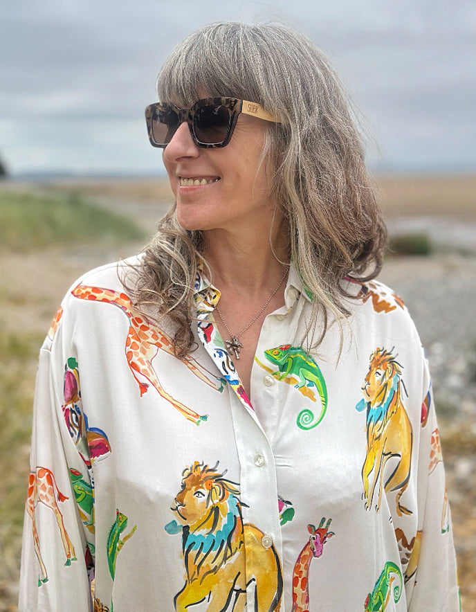 A woman with gray hair and sunglasses wearing a white button-down shirt with a colorful safari animal print of lions, giraffes, and chameleons, set against a blurred outdoor background.