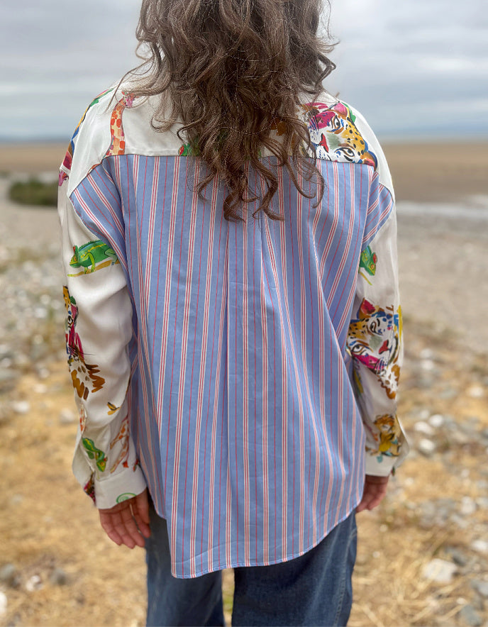 A person viewed from behind wearing a long-sleeve button-up shirt with a blue, orange, and white vertical stripe body and multicolored floral-patterned sleeves, standing outdoors on a rocky beach.