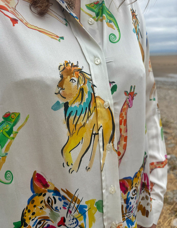 A close-up of a white button-up shirt featuring a colorful safari animal print with a prominent yellow lion, green chameleon, and giraffe, worn by a model outdoors.